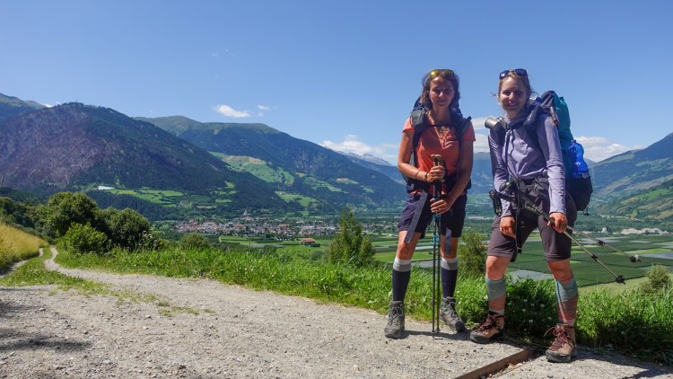 Zwei Wanderer stehen auf einem Wanderweg in den Bergen, umgeben von grünen Wiesen und Bergen im Hintergrund, mit einem Dorf in der Talsohle.