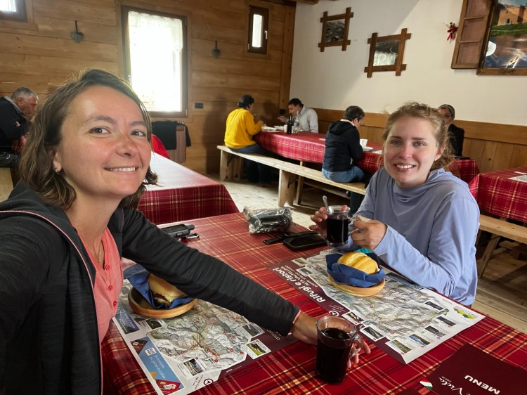 Zwei Frauen lächeln in einem rustikal eingerichteten Restaurant, während sie eine Mappe und Essen auf einem karierten Tisch decken. Ein Teil des Raumes mit Gästen im Hintergrund ist sichtbar.
