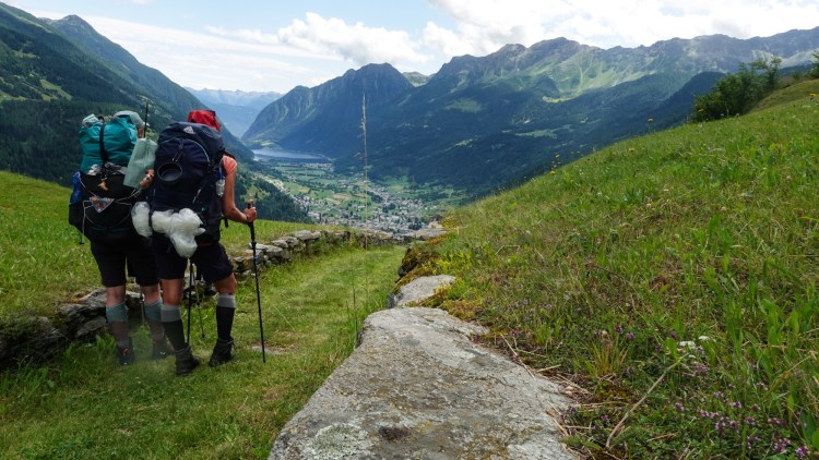 Zwei Wanderer mit Rucksäcken stehen auf einem Wanderweg und blicken auf ein Tal mit Bergen im Hintergrund.